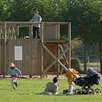 Ein Mann liegt neben einem Kinderwagen auf einer Wiese. Im Hintergrund spielen Kinder auf dem großen Abenteuerspielplatz aus Holz..