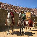 Two gladiators riding into the arena in front of packed stands, followed by actors holding a banner.