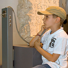 Junge hält sein Ohr an eine Hörstation vor einem Stein mit römischer Inschrift.