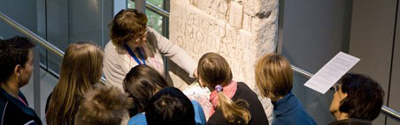 Eine Führerin erklärt einer Schülergruppe die Inschrift eines römischen Grabsteins." 