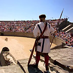 Ein Mann in römischer Kleidung schaut auf das große, mit zahlreichen Zuschauern gefüllte Amphitheater.