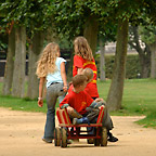 Auf einer Parkallee ziehen zwei Mädchen zwei andere Kinder im Bollerwagen hinter sich her.