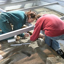 Zwei Menschen arbeiten für die Ausstellung im RömerMuseum an einer Fläche, auf der sich römerzeitliche Laufspuren erhalten haben.