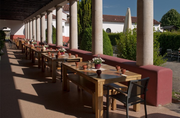 Perspektivischer Blick auf gedeckte Tischreihen in einem Säulengang, dahinter der gepflegte Garten der römischen Herberge