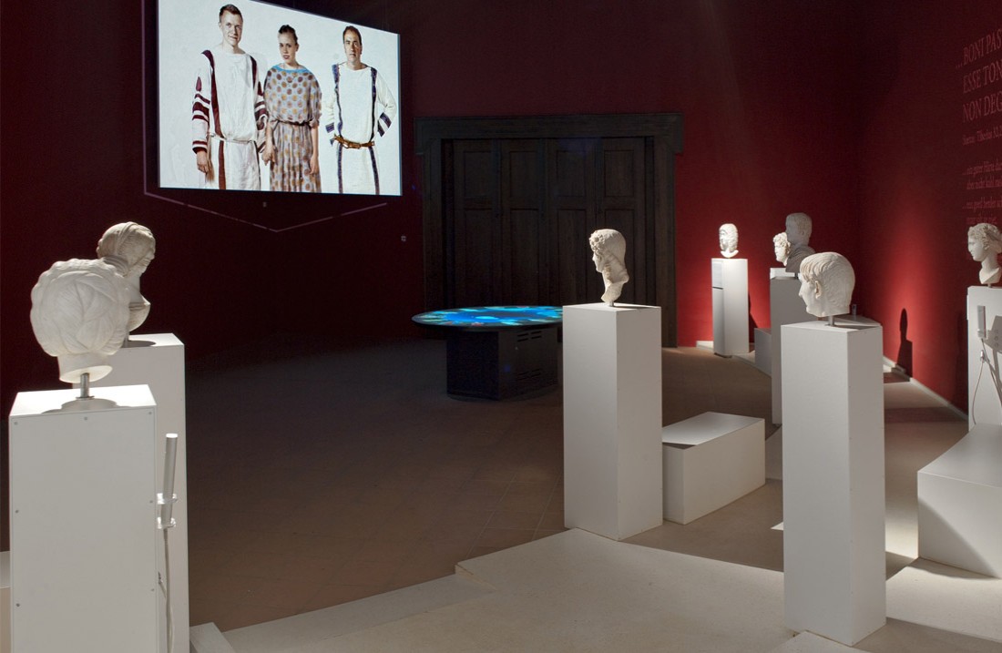 Roman statues and busts on pedestals, film presentations in the background