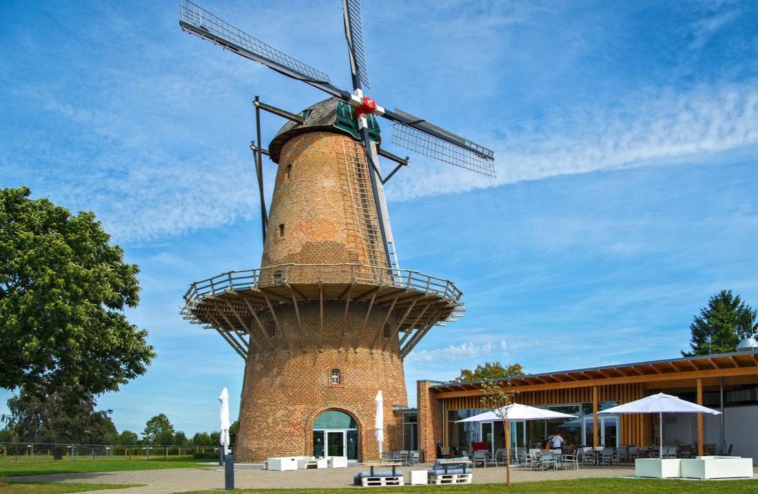 Windmolen met rechts ervan een plat gebouw, ervoor tafels en stoelen