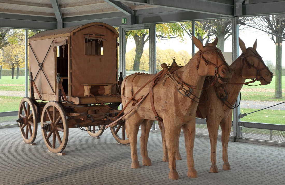 Ein kleiner, hölzerner vierrädriger Planwagen mit hölzernen Pferdefiguren in einem Pavillon aus Glas und Stahlträgern