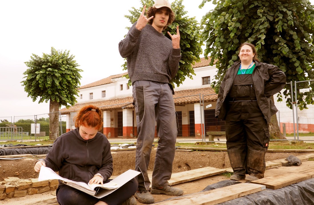Drie studenten van de IASX poseren aan de rand van een opgraving, met op de achtergrond de ambachtsliedenhuizen van de APX