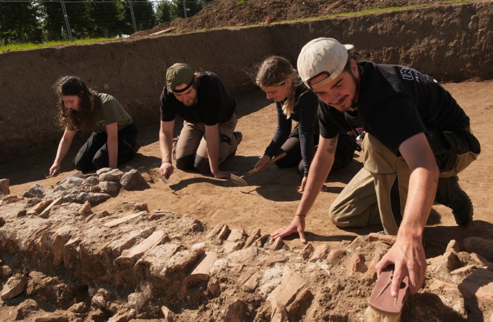 Vier junge Archäolog:innen bei der Ausgrabung, in der Hocke vor einem Stück Grundmauer