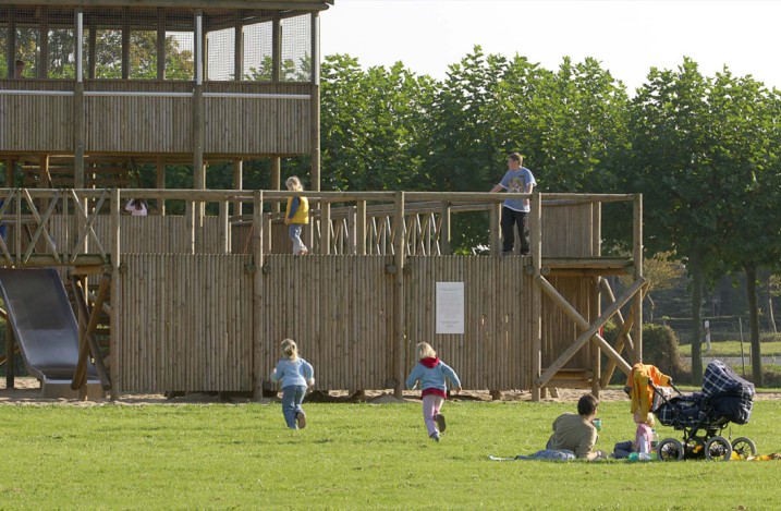 Grote houten speeltuin van twee verdiepingen met palissaden en torens, veel spelende kinderen, verschillende ouders met kinderwagens picknicken op het grasveld ervoor 