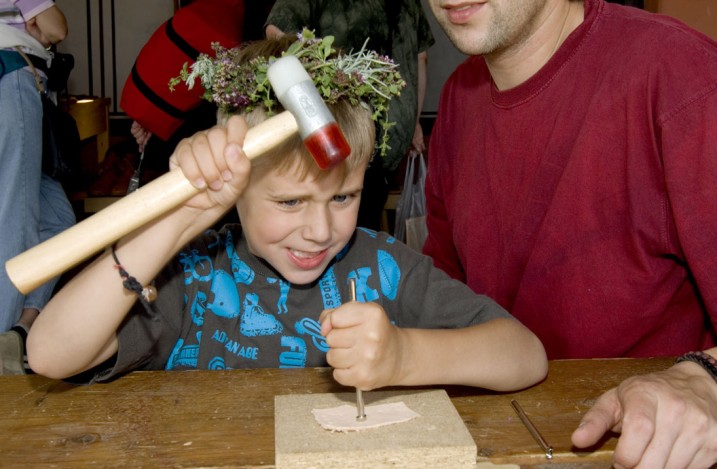 Ein Kind von etwa 7 Jahren punziert ein Stück Leder, im Anschnitt sieht man einen jungen Mann, der auf das Kind aufpasst