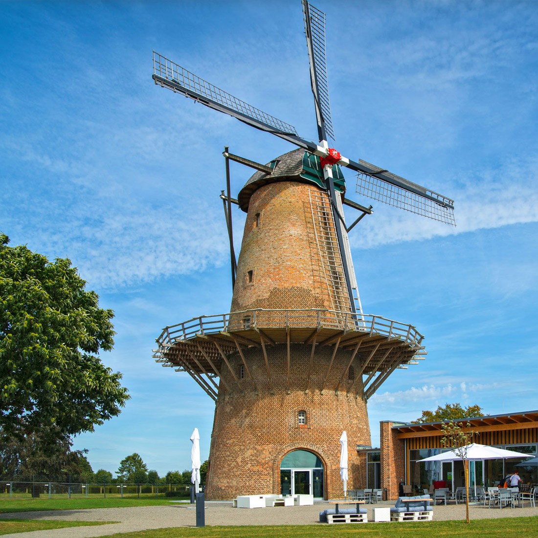 Windmühle im Park mit einem flachen, modernen Gebäude rechts daneben, davor Stühle, Tische und Sonnenschirme